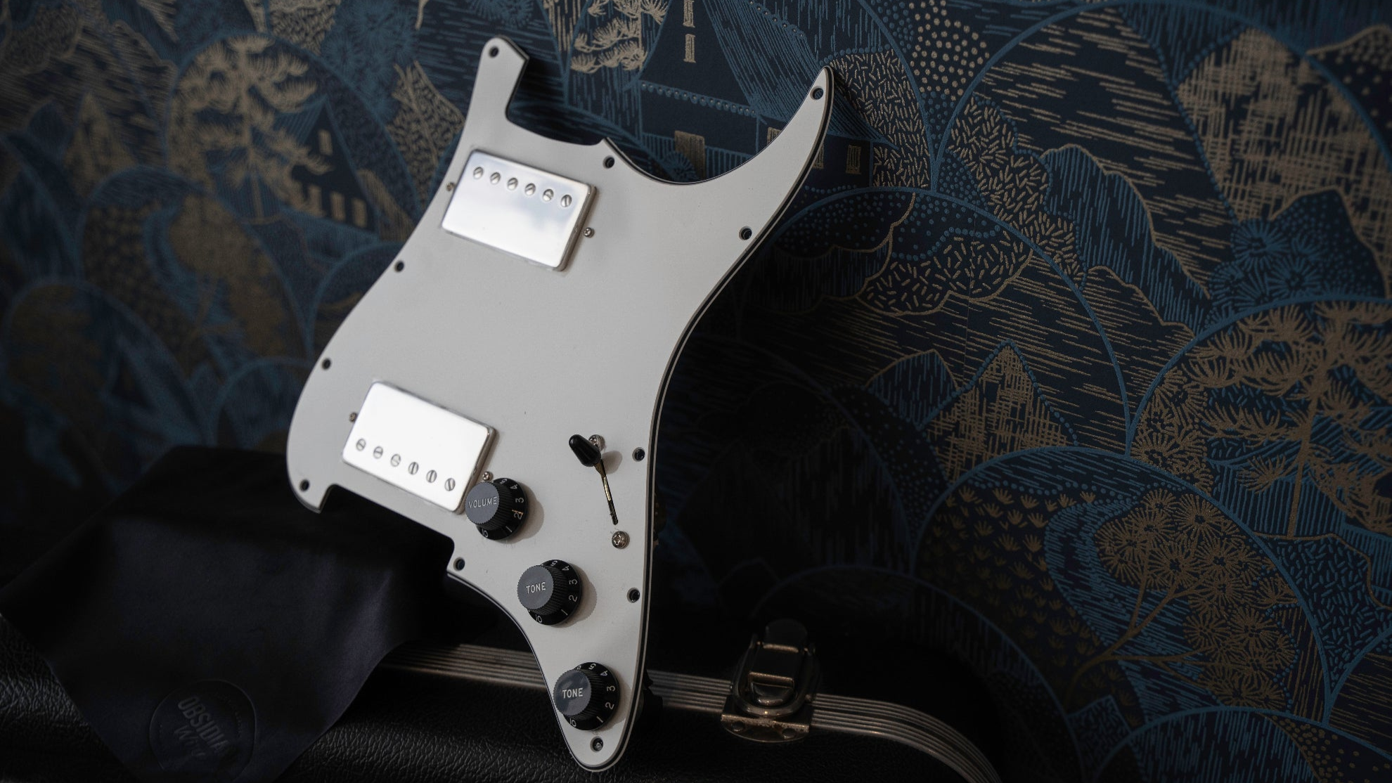 a parchment white loaded pickguard for HH Stratocaster is placed on top of a guitar case. The pickguard is rested against a wall with patterned blue and gold wall paper.