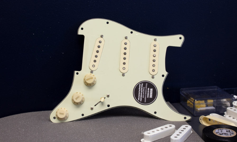 A mint green SSS loaded pickguard with aged white knobs and pickup covers is rested against a wall, with pickguard building supplies in the foreground.
